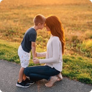 Mother and son looking into each others eyes and heads touching. Low sunlight creating a peaceful setting. Mother has long brown hair, light skin and white top. Child has short blonde hair, light skin blue teeshirt.