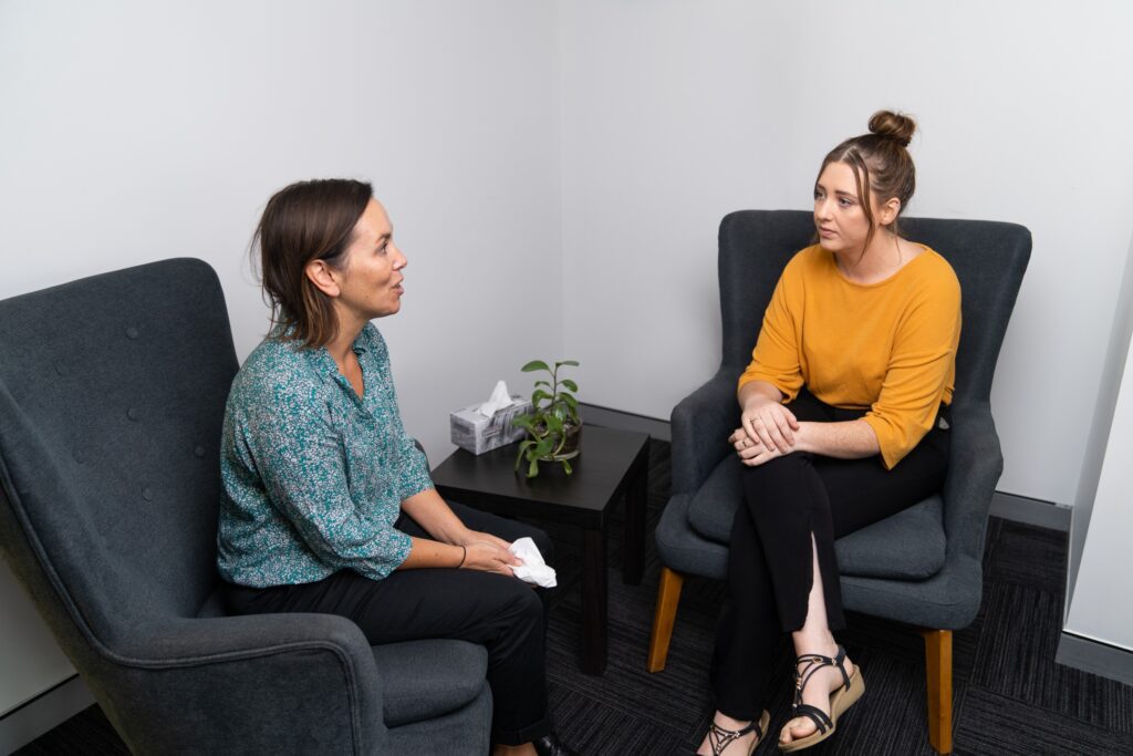 Two women in sitting and talking in the CADA counselling room. One women is siting, listing and understanding, while the other is talking and holding a tissue.
