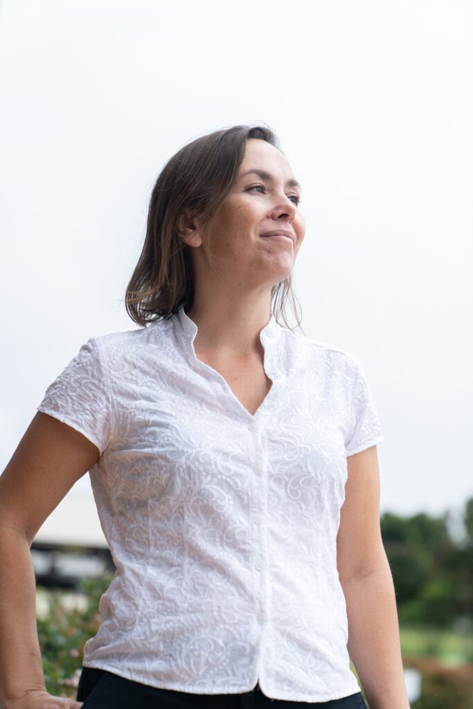 Woman standing tall, looking content and empowered. She is wearing a white top and has short brown hair and light skin.
