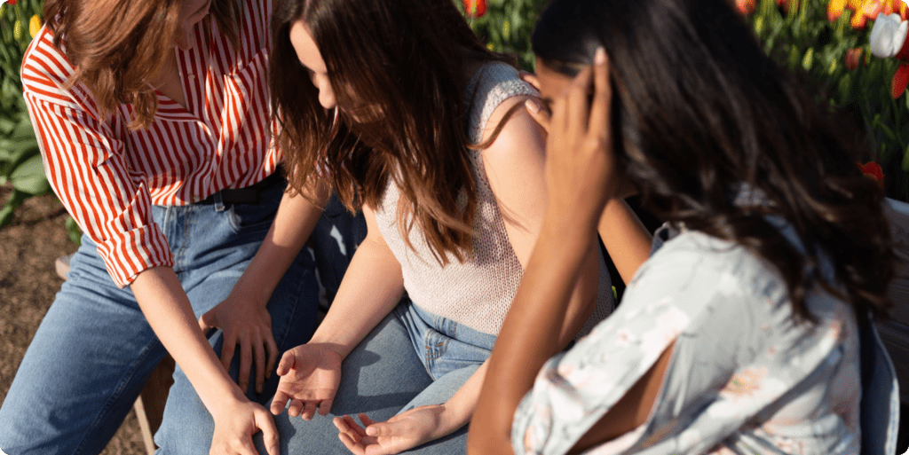 Three young women sitting on a bench. Women in the middle is talking to her friends while they consolidate her.