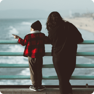 Mother and child from behind looking over a railing at the ocean. Mother is wearing a black jacket and child is wearing a red and white checkered jacket with a black beanie. Child is pointing out to sea.