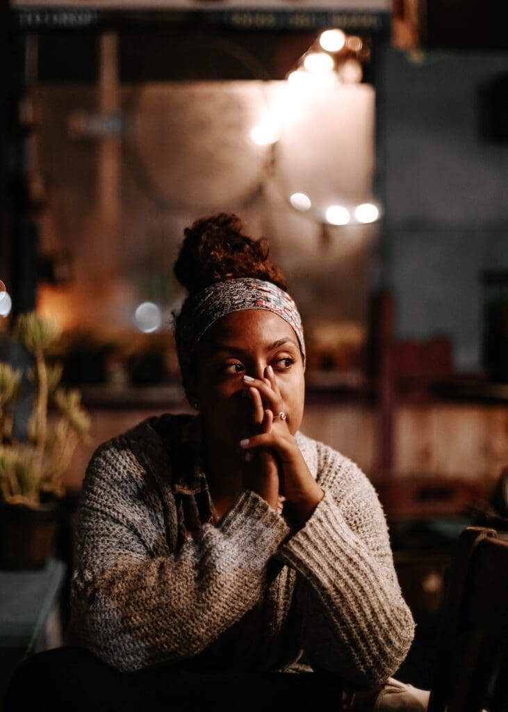 Pensive woman sitting and reflecting with hands covering mouth. Woman is wearing a light brown jumper and multi-coloured headband, she has dark brown hair tied in a bun and dark skin.