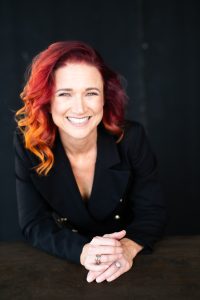A portrait of a smiling woman with red hair against a black backdrop.